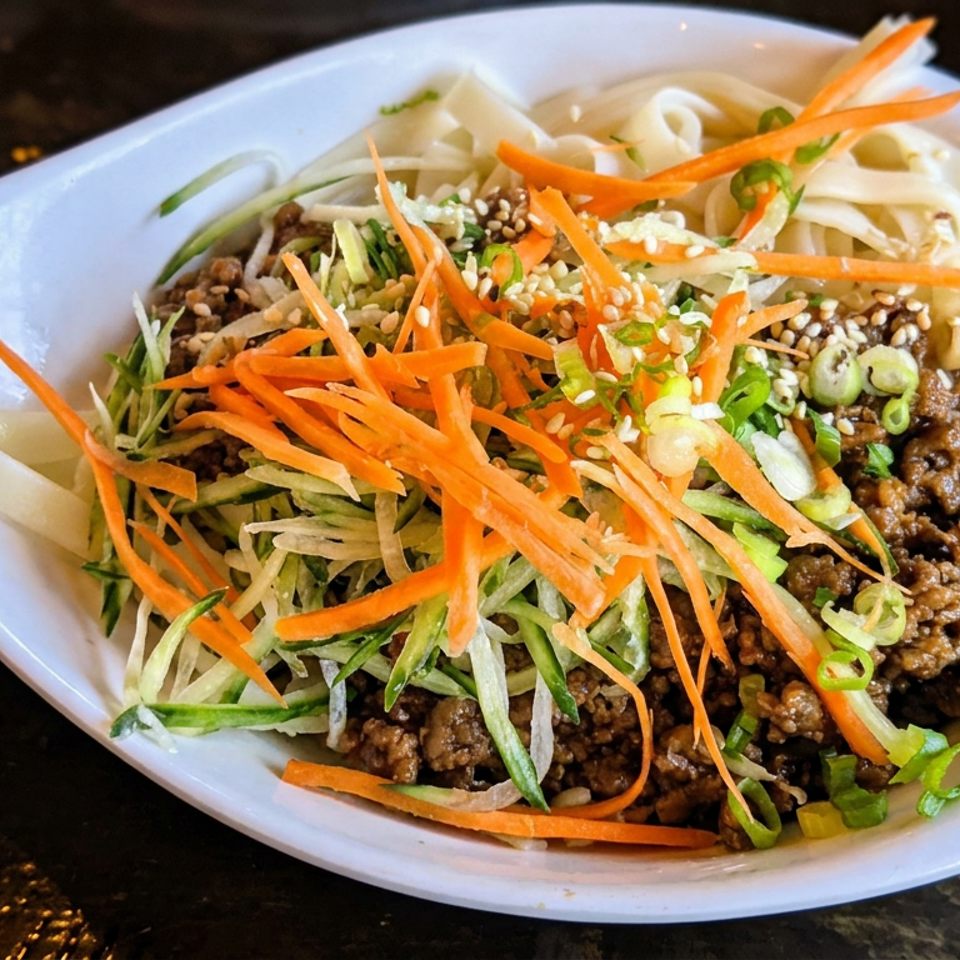 Bowl of noodles topped with seasoned ground meat, shredded carrots, cucumber, scallions, and sesame seeds.