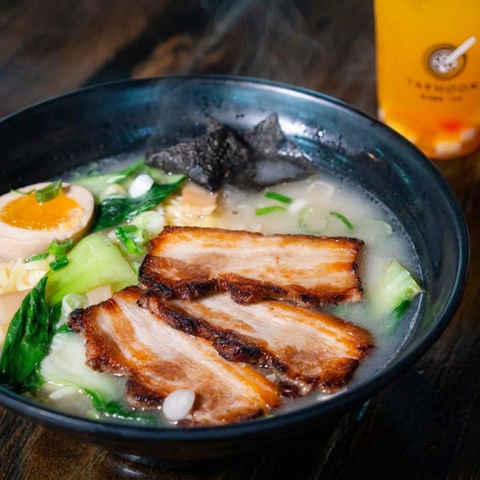 Bowl of ramen with sliced pork belly, soft-boiled egg, greens, noodles, and seaweed beside an orange drink on a wooden table.