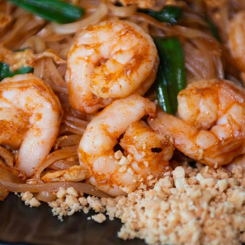 Close-up of seasoned shrimp and translucent noodles with green onions and crumbled topping on a dark plate.